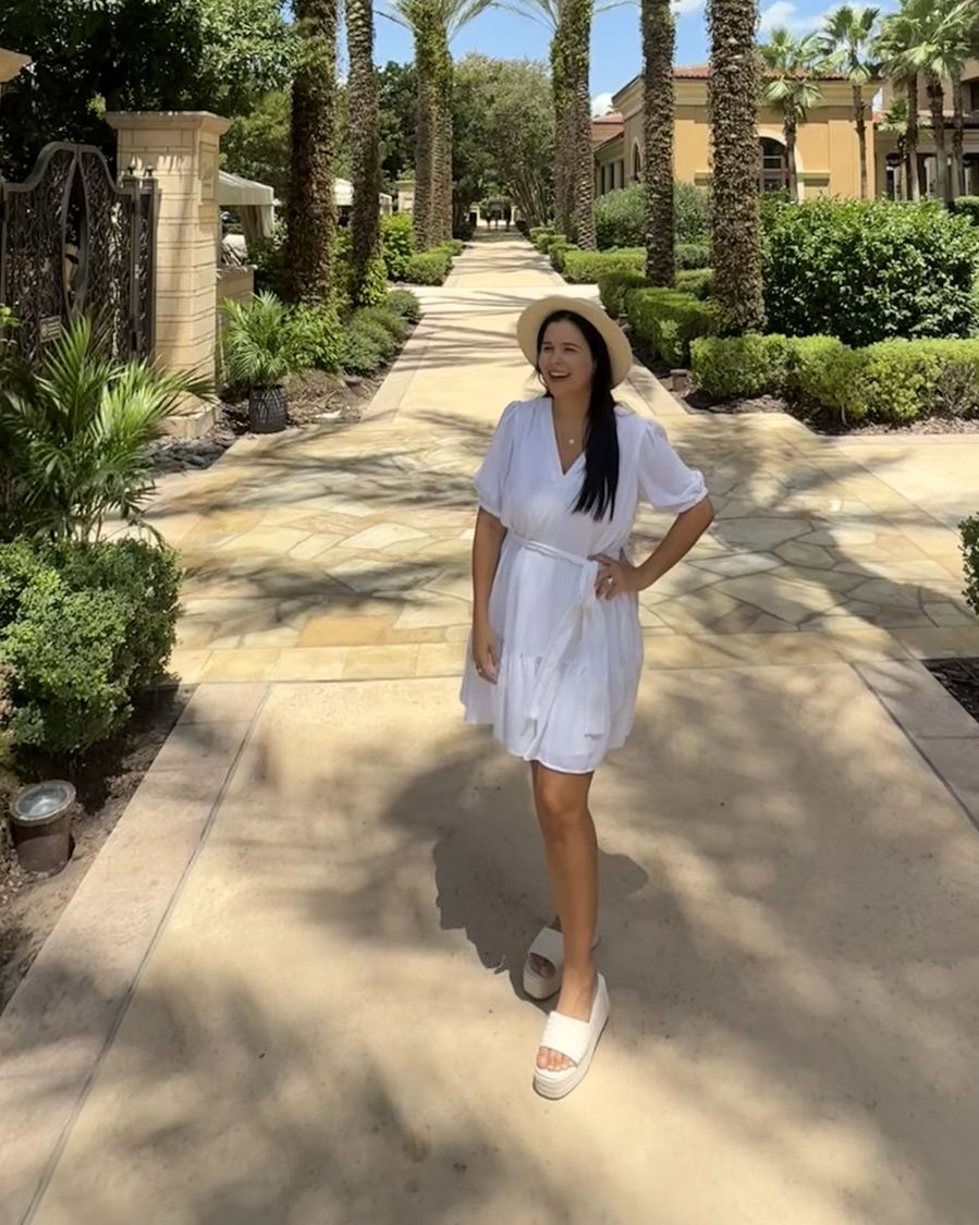 Rafaela in a white dress and sun hat on a palm-lined walkway